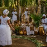 Espaço ao ar livre com altar decorado, pessoas vestidas de branco realizando cerimônia espiritual em meio a plantas tropicais.